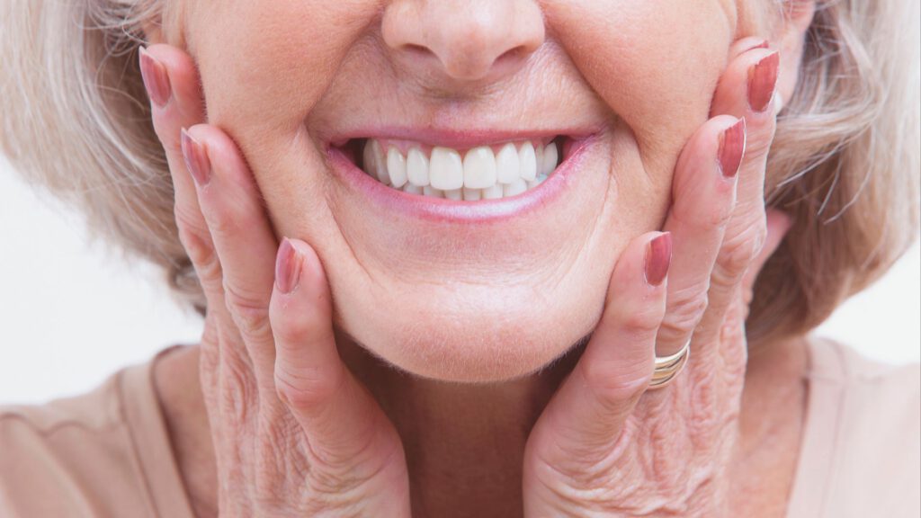Eine ältere Frau mit strahlendem Lächeln, die ihre Wangen berührt und ihre perfekten Zahnersatz zeigt.
